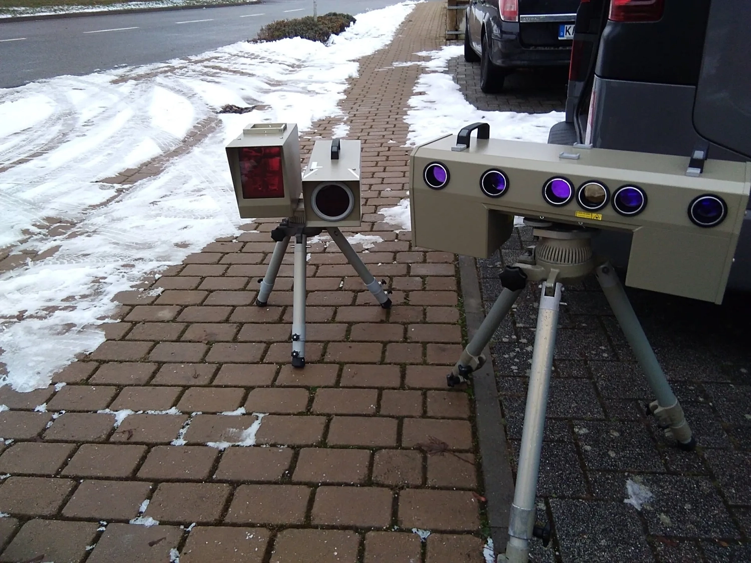 Radarrent GmbH – Verkehrsüberwachung – Zwei auf Stativen montierte Radargeschwindigkeitsmessgeräte auf einem schneebedeckten Backsteinpflasterweg in der Nähe geparkter Autos